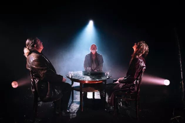 Three persons sitting around a table in theatre scene. Photo.